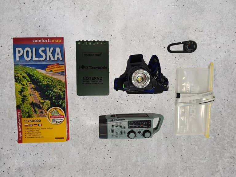 Moduł Orientacja rozłożony na betonie: mapa samochodowa Polski, notes wodoodporny, radio awaryjne z korbką i panelem solarnym, latarka czołowa, etui na dokumenty i karabińczyk z kompasem.